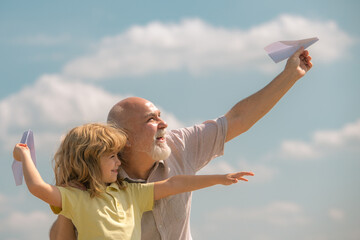 Old grandfather and young child grandson with paper plane over blue sky and clouds. Men generation granddad and grandchild.