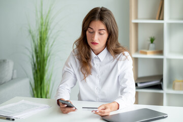 Online payment. Office woman. Mobile application. Concentrated elegant lady holding credit card typing smartphone sitting work desk in light room interior.
