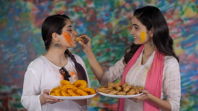 Young and beautiful Indian sisters celebrating Holi and offering sweets to each other. Smiling females with local Indian sweets - Imarti and Gujiya  Festival of Holi