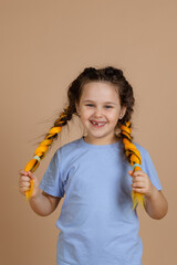 Excited, happy cheerful little girl smiling with missing tooth looking at camera holding kanekalon braids with hands on beige background wearing blue t-shirt and jeans.
