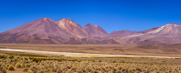 Panorama image of arid altiplano landscape with multicolored volcanoes and dry salt lake under blue...