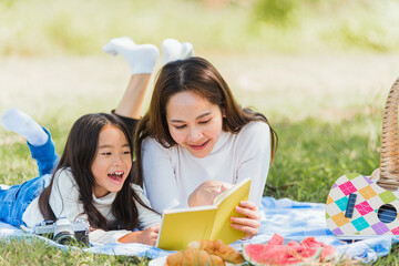 Happy Asian young family mother and child little girl having fun and enjoying outdoor laying on picnic blanket reading book at summer garden spring park, Family relaxation concept