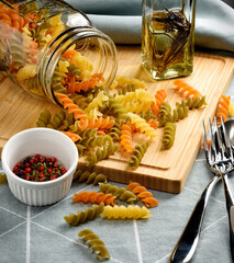 Tricolor dry pasta, rotini, fusilli pour from glass jar on wooden board with red and black pepper in white bowl in foreground and rosemary olive oil in glass bottle in background