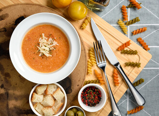 tomato soup garnished with pepper, parsley and parmesan cheese in white bowl on wooden board with bread cube and red black pepper and tricolor rotini fusilli on side, top view, flat lay