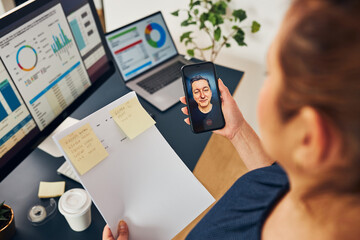 Businesswoman having video chat on mobile phone with her colleague. Businesswoman working with data on charts, graphs and diagrams on computer screen. Woman holding smartphone sitting in office