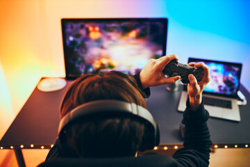 Man playing video game at home. Gamer holding gamepads sitting at front of screen. Streamer playing online in dark room lit by neon lights. Competition and having fun