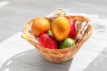 A basket to multicolored Easter eggs, feathers and a willow branch on a white wooden background with shadows.