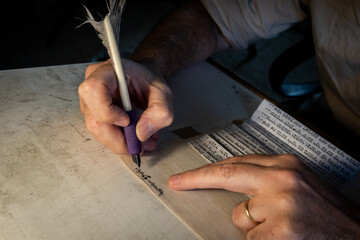 Closeup view of the hands of a Jewish scribe holding a feather quill and writing the words of the...