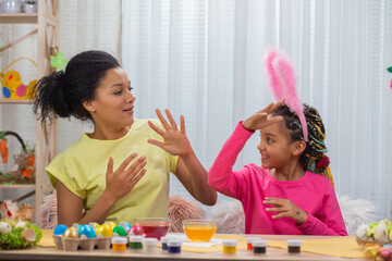 Mom and daughter with funny bunny ears funny dance and having fun. African American woman and little girl are sitting at table in festively decorated room at home. Happy easter. Close up.