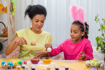 Mom and daughter with funny bunny ears dips eggs in food coloring using yellow and red paint. African American woman and little girl are sitting in decorated room. Happy easter. Close up.