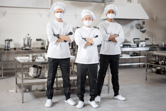 Full body portrait of multiracial team of three chefs standing together in the professional kitchen. Well-dressed chefs in face masks and protective gloves ready for a job. New normal for business