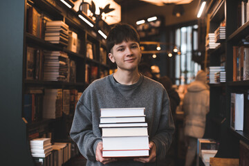 Smiling teenager boy 17-18 year old holding stack of paper books standing in public library indoor....
