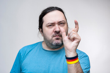 An adult, unshaven man in a T-shirt shows a small size with his fingers. Bracelet in the colors of the German flag.