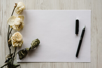 Composition dried rose flowers, sheet of paper and pen for message on a white wooden background. Postcard with copy space. Frame. Place for text, design. Greeting card. Flat lay, top view.
