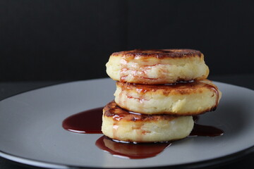 food breakfast cheesecakes curd pancakes three pieces lie stack of slides on top of each other and pour jam sauce cherry red on a gray plate on a black background