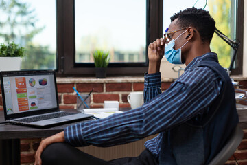 Uninspired entrepreneur wearing face mask feeling tired looking at laptop screen with business analytics sitting at desk. Startup owner with covid protection thinking about sales problems.