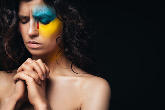 A Ukrainian carly girl with a bloody tear prays for peace in Ukraine. Make-up in the form of the ensign of Ukraine. War in Ukraine.