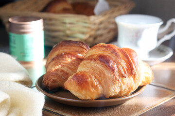 A cooked French croissant in a  plate with coffee and wooden basket in background. Sun light from window in the morning. It is a buttery, flaky pastry. Made with a yeast leavened dough 