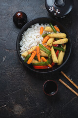 Black bowl with panasian stir-fried vegetables and white rice, top view on a dark-brown stone background, vertical shot with space