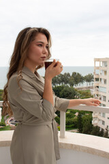 beautiful asian girl is standing on a balcony and drinking a coffee, sea is on background