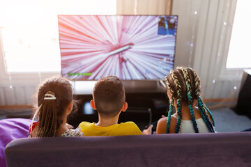 little children a boy and two girls with pigtails play a game console in front of a big TV in the playroom with their backs turned