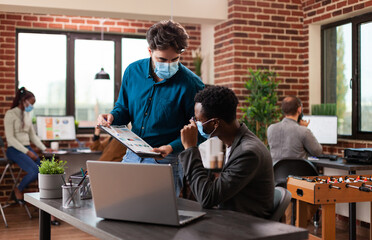 Diverse entrepreneurs working at company collaboration analyzing marketing graph discussing business ideas in brick wall startup office. Multi-ethnic team wearing medical face mask against covid19