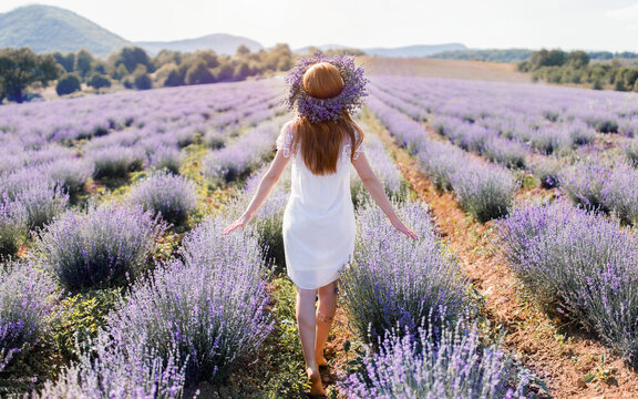 a barefoot young girl in a white light dress with long red hair and a wreath on her head walks along a large lavender field among the mountains with her back turned