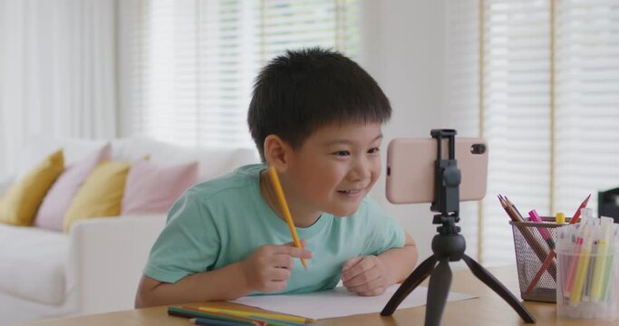 Asia cute smart boy alpha gen z kid enjoy fun hobby idea paint pencil color game on paper at online day care play school by vlog talk video call app. Young son study class tutor teach at home table.
