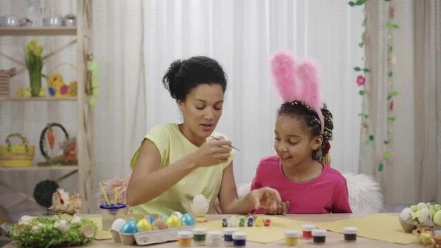 Mom teaches her daughter to paint eggs with paints and brush. African American woman and little girl with bunny ears are sitting at table in decorated room. Happy easter. Slow motion ready 59.97fps.