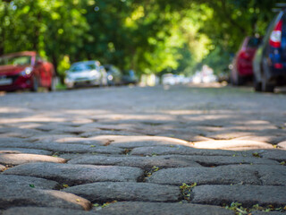 A city alley paved with cobblestones and parked cars in the shade of trees in a quiet, peaceful area. Focus on the foreground. A European paved street lined with cars