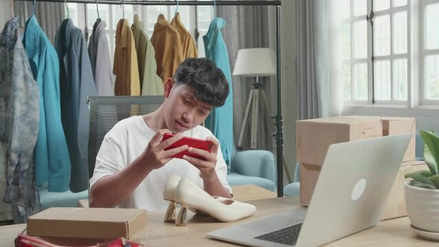 Asian Man Online Seller Taking Photos With Mobile Phone While Using Computer For Selling Clothes At Home
