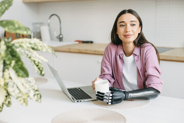 Cute adorable female of 20s with long lashes and sensual lips sitting at table with white cup of coffee in hands, one arm made of metal bionic mechanical prosthesis after amputation