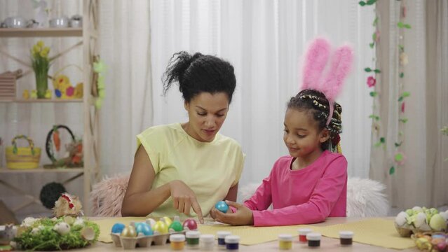 Mom and daughter with funny bunny ears gluing pictures on colored eggs. African American woman and little girl are sitting in festively decorated room at home. Happy easter. Slow motion ready 59.97fps