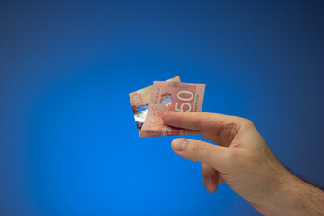 Paper banknote of 50 Canadian dollar bill held between fingers by Caucasian male hand. Close up studio shot, isolated on blue background