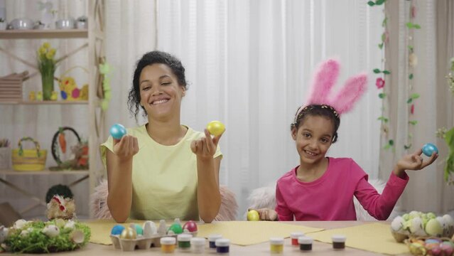 Mom and daughter with funny bunny ears laugh and have fun with colored eggs. African American woman and little girl sitting at table in decorated room at home. Happy easter. Slow motion ready 59.97fps