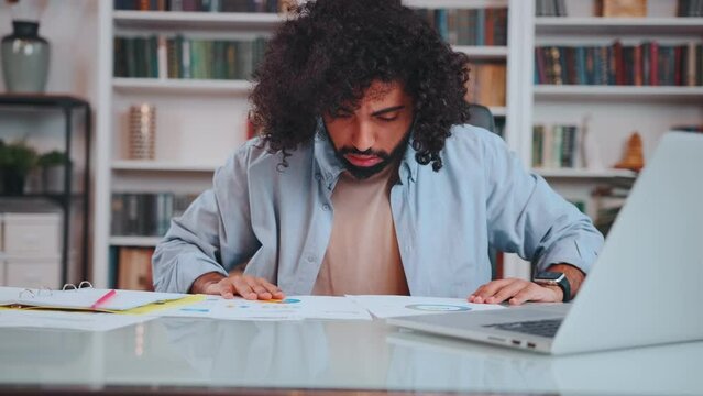 Young perplexed Middle Eastern man financier nervous looking through analytical reports demonstrating indicators of crisis and recession sits at table with laptop. Financial analysis, forecasting
