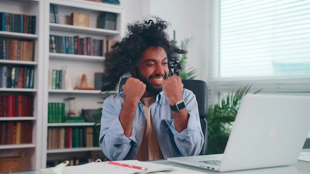 Young excitable Arabian American man screams with joy and waves hands looks at laptop screen after winning online casino and getting jackpot sits at table in home office. Gambling addiction