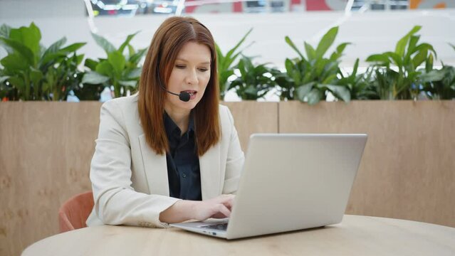 Businesswoman wearing headset talking to webcam and typing on laptop, tracking shot. Online consultant giving support. Concept of technology