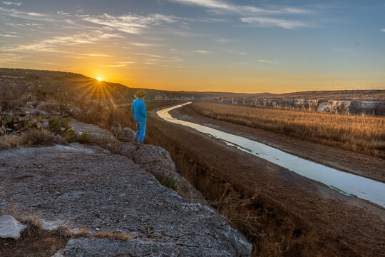 Person, man, wearing a bike helmet, standing on the edge of a cliff, watching the sunrise over the Rio Grande river flowing below. Seminole State Park, Texas