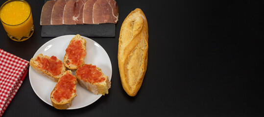 Tostadas con tomate natural, tapa de jamón ibérico, y zumo de naranja, todo dispuesto sobre mesa de madera de color negro