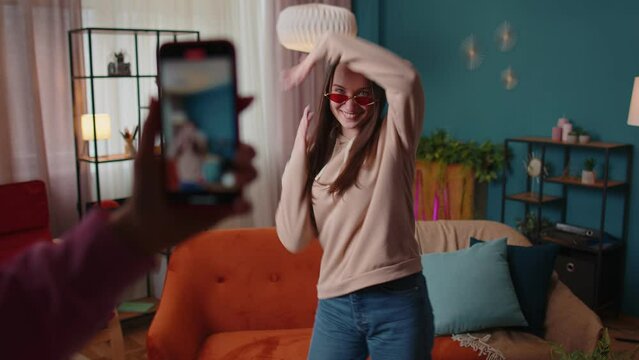 Blogger Concept. Joyful teen girl dancing at camera filming video using mobile phone at home in living room. Young woman creating her trendy content on a smartphone app to share on social media