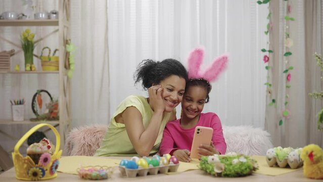 Mom and daughter with funny bunny ears take selfies and have fun. African American woman and little girl are sitting at table in festively decorated room. Happy easter. Slow motion ready 59.97fps.