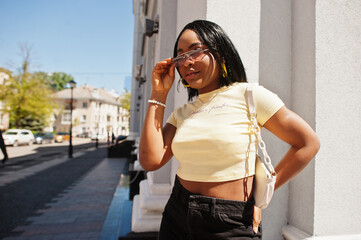 Fototapeta premium Beautiful fashionable young african american woman posing in street, sunglasses, yellow top. Fashion summer photo. Bright colors. Nice view afro people.