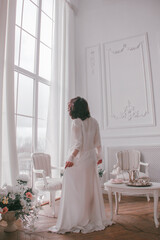 young girl with black short hair in white boudoir robe is standing from the back near huge panoramic window on the white wall background with flowers. lifestyle concept, free space