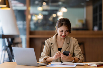 Photo of smiling asian woman using cellphone while working with laptop in office