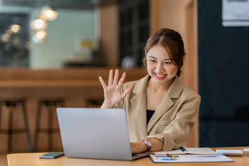 Fototapeta na wymiar Joyful young woman sit at the work desk, waves hand, greeting colleagues. Attractive business female communicate with colleagues or friends by video call, smiling, remotely work or study