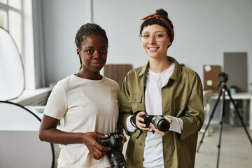 Waist up portrait of two female photographers smiling at camera while standing in photo studio,...