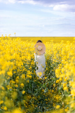 woman in a long white dress and a big hat runs through a rapeseed field. Photo from the back in yellow flowers on a blue sky background