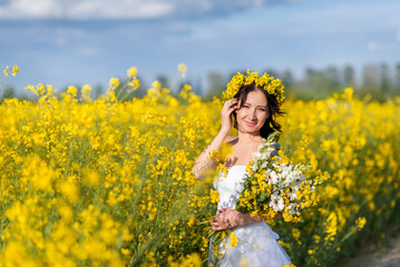 Naklejka na ściany i meble Portrait of a beautiful woman with a wreath of yellow flowers on her head. A girl with a bouquet in a rapeseed field