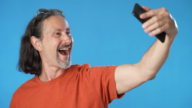 Happy elderly bearded mature man 50s 60s orange t-shirt doing selfie with mobile phone post photo on social network isolated on solid blue background copy space